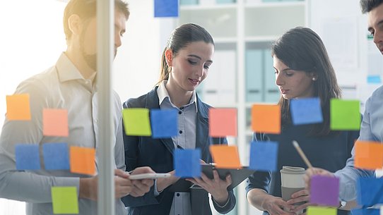 Arbeitsgruppe steht vor Glasscheibe mit Post-Its. 